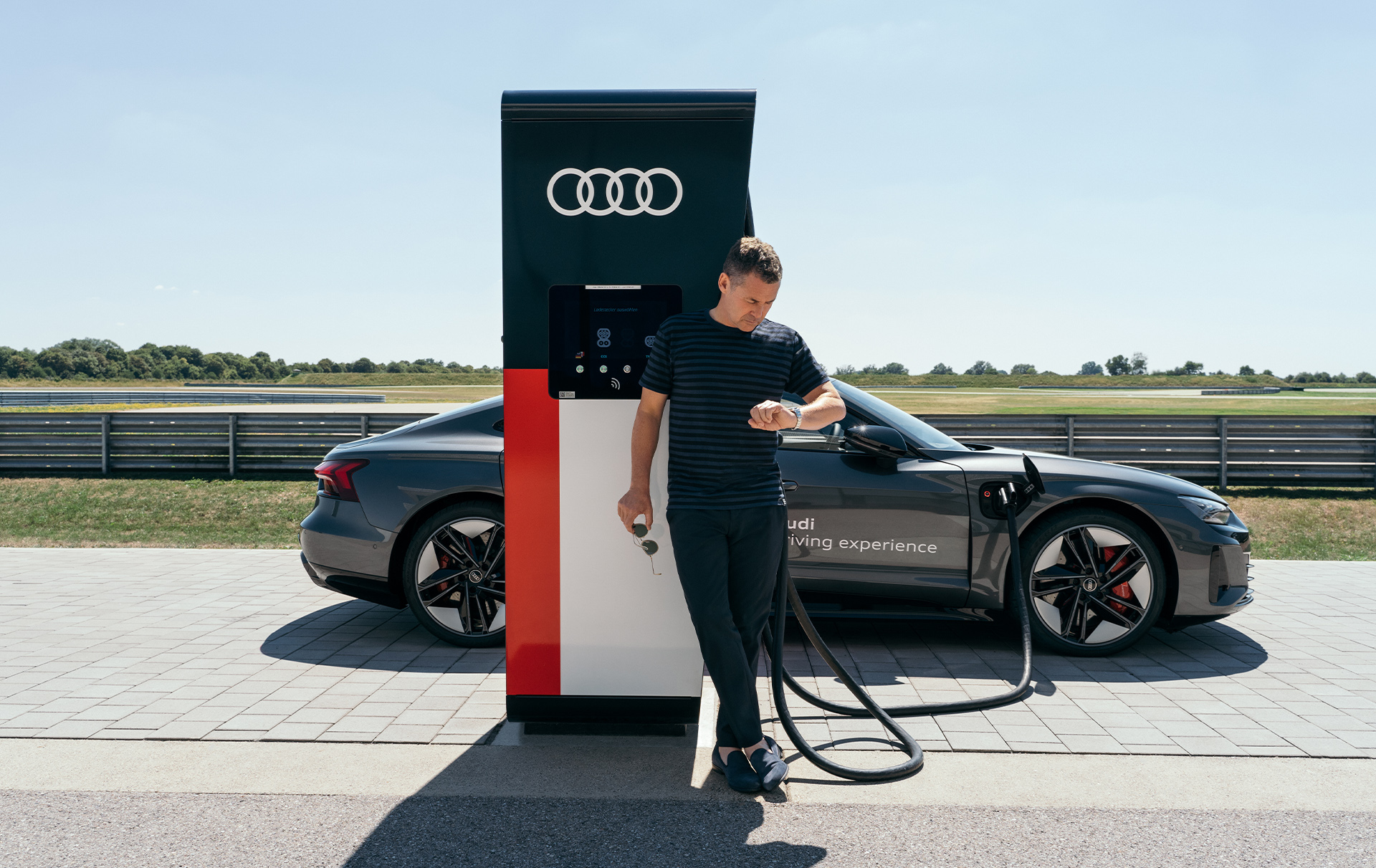 Tom Kristensen kijkt op zijn horloge terwijl de Audi RS e-tron GT aan de snellader hangt.
